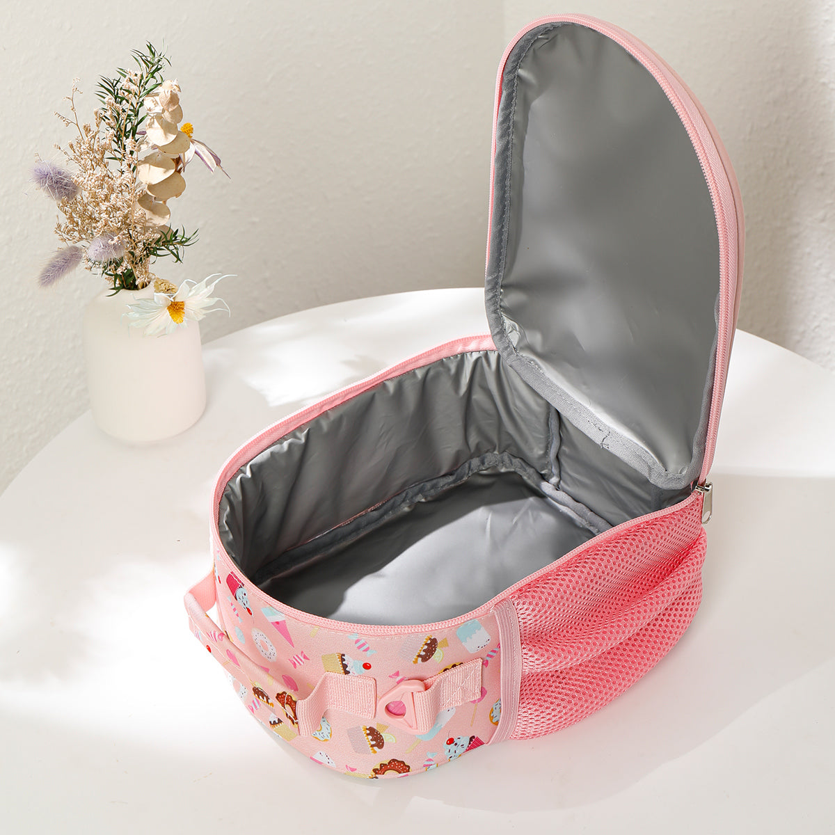 Pink insulated lunch bag with floral design on a white surface with a vase of flowers in the background.